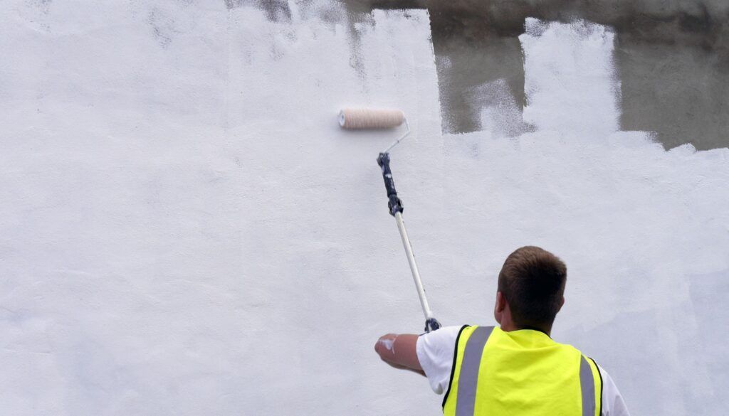 Person in a yellow vest painting a wall white with a roller.