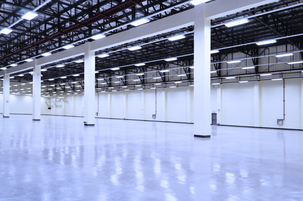 Large, empty, brightly lit warehouse with polished concrete floor, white walls, and exposed ceiling beams.