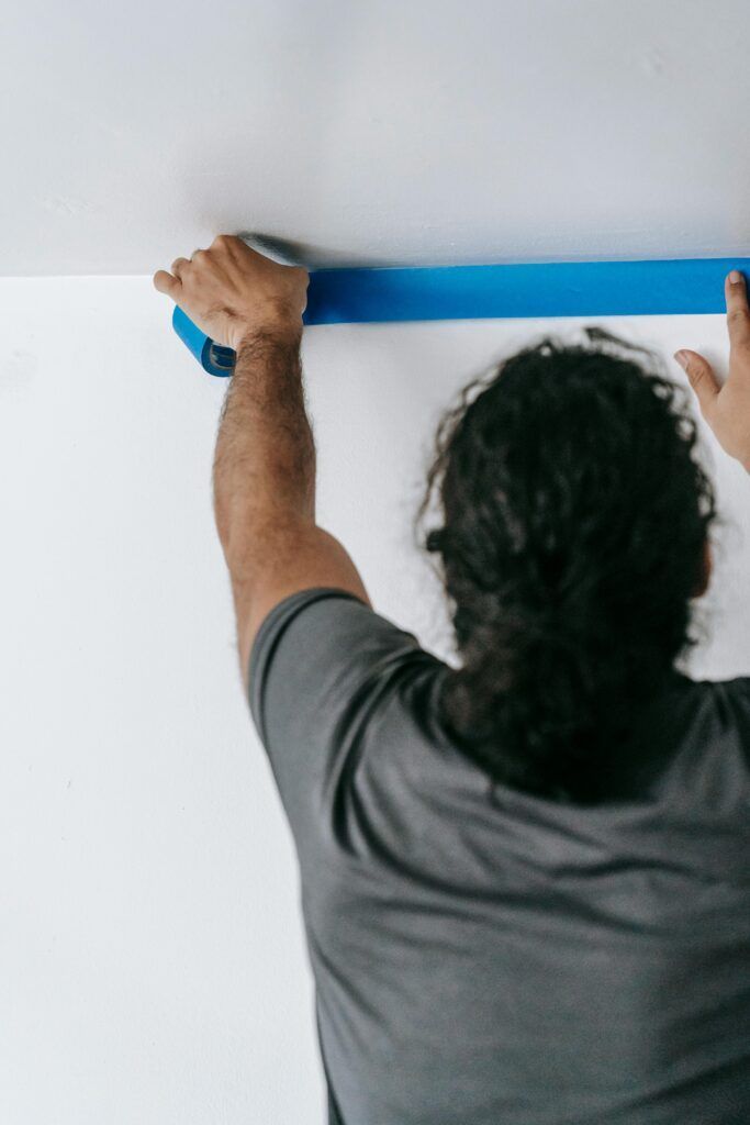 Person taping the wall/ceiling with blue painter's tape, preparing for painting.