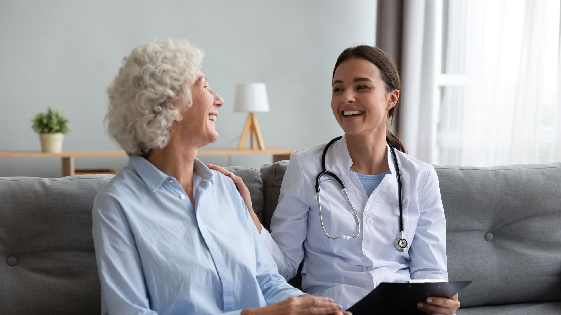 Doctor and elderly patient smiling on a gray couch in a bright living room, with a stethoscope visible.