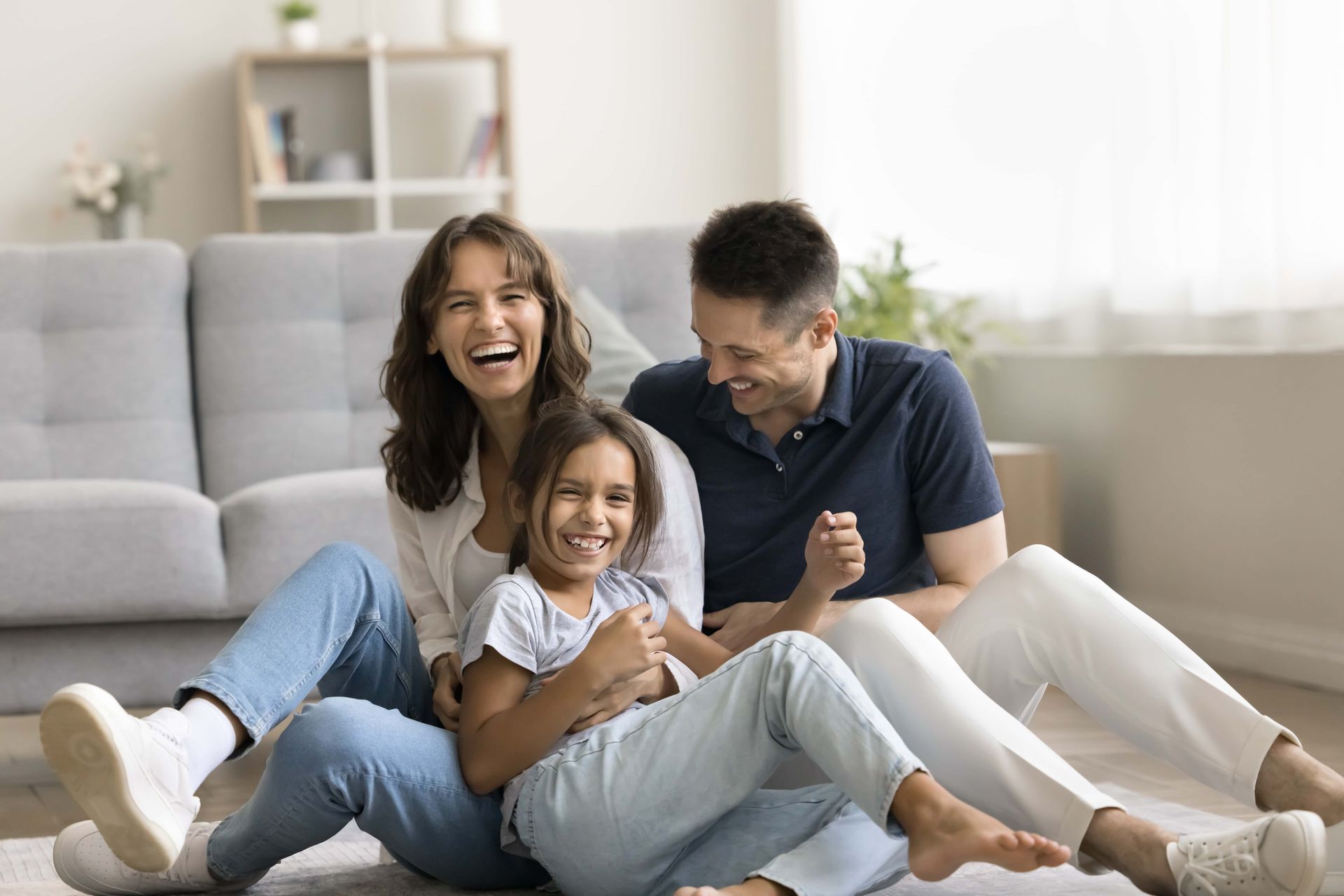 Three people laughing and sitting on the floor in a bright living room, with a gray sofa behind them