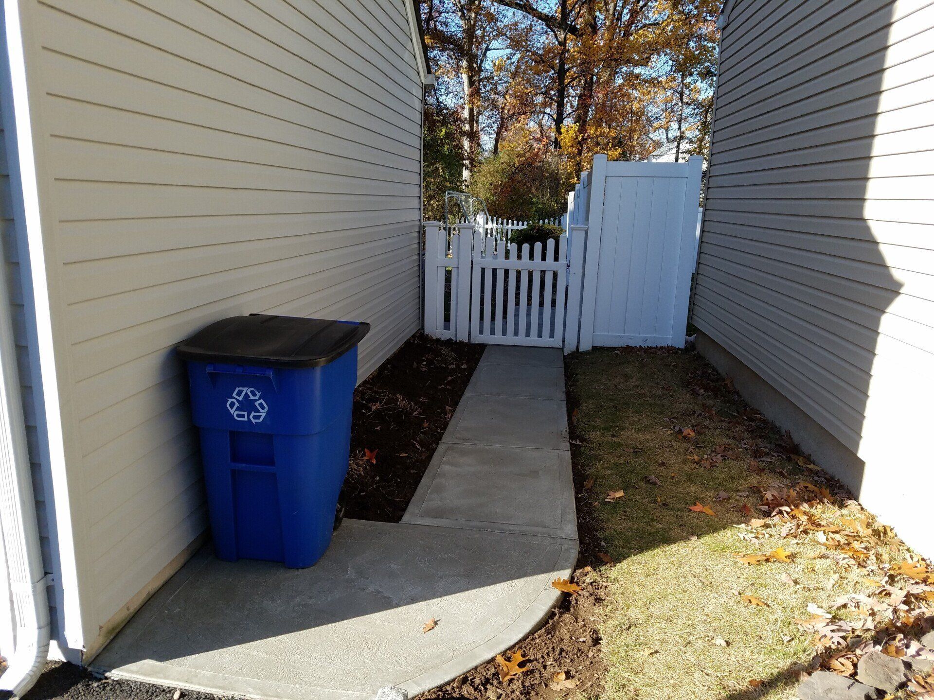 A blue trash can is sitting on the side of a house next to a walkway.