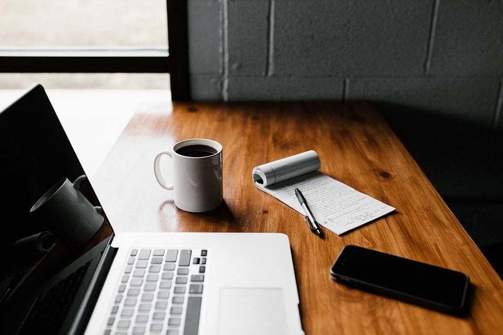 Laptop, coffee mug, notepad, and smartphone on a wooden desk next to a window.