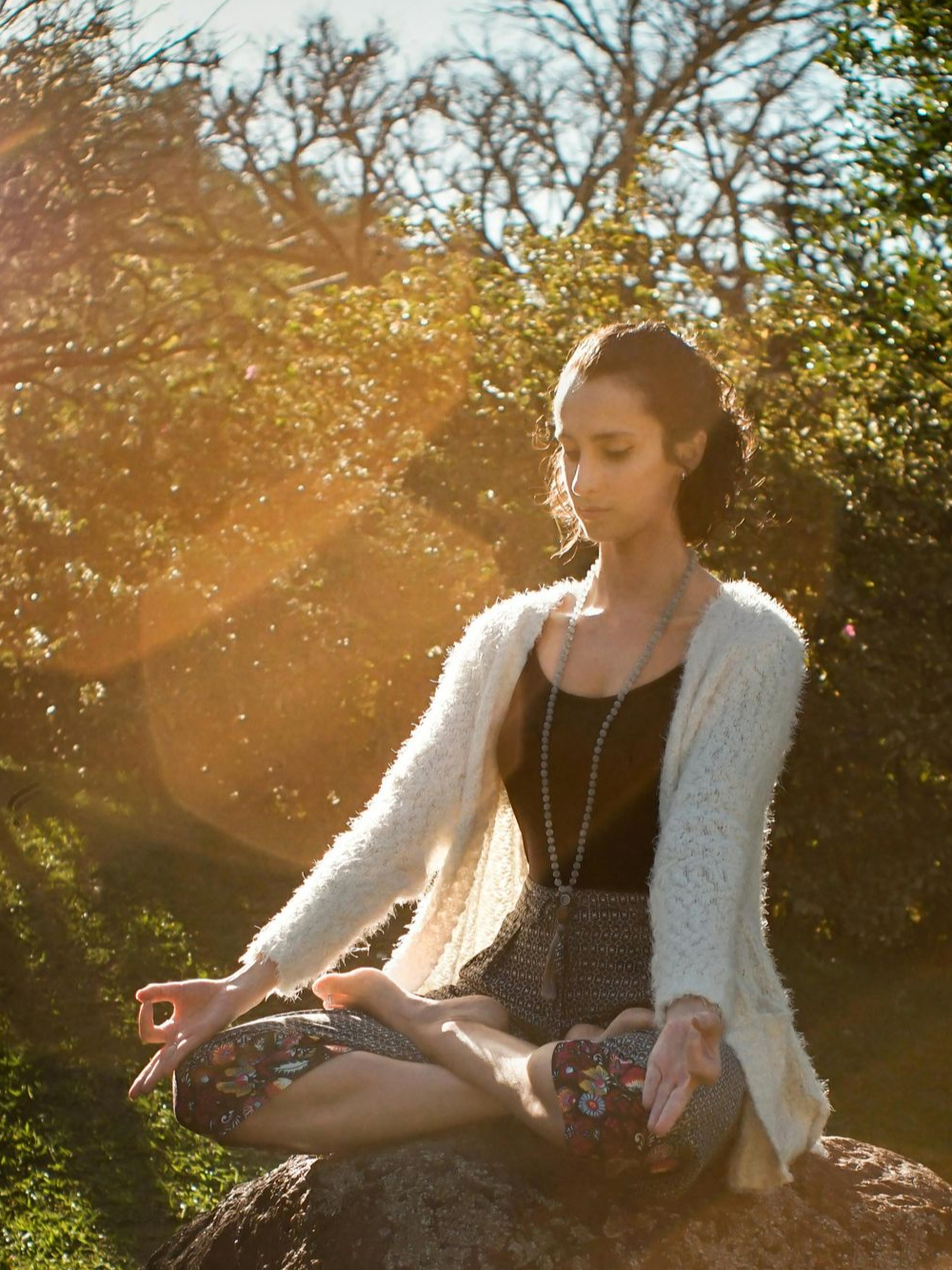 Mujer meditando al aire libre, a la luz del sol, sentada con las piernas cruzadas sobre una roca.