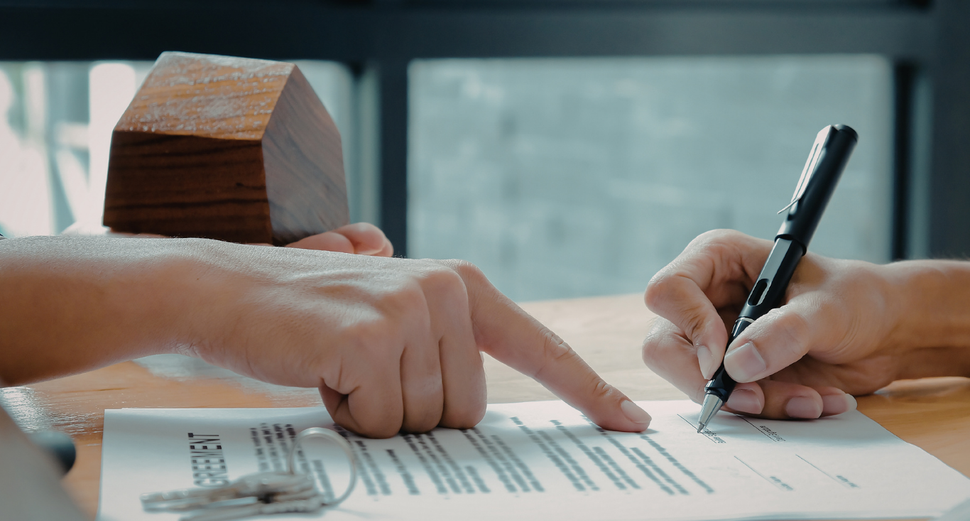 Two hands over a document: one points at text while the other prepares to sign, with a small wooden house model nearby.