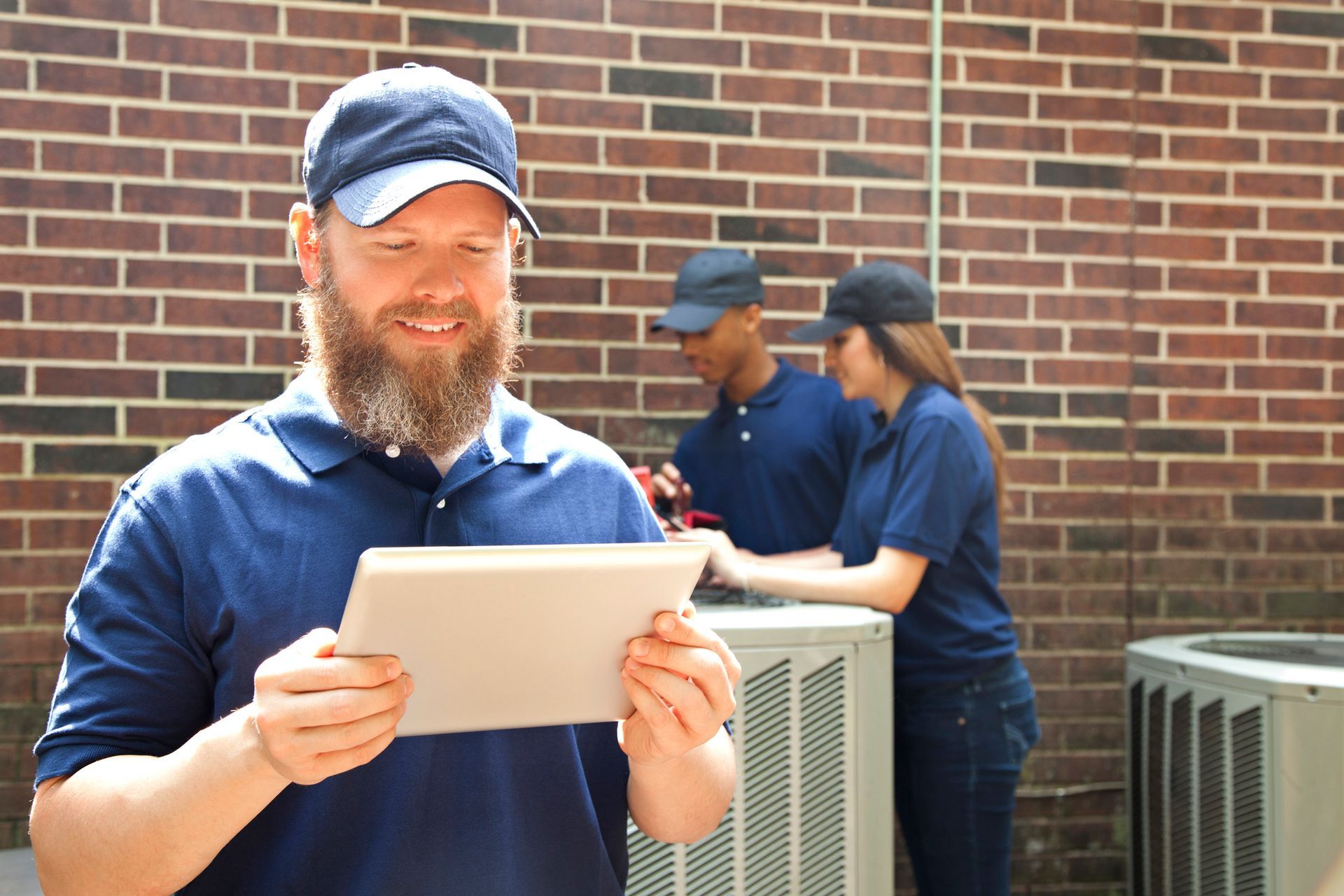 HVAC technician with tablet, smiling, while two colleagues work on AC unit against a brick wall.