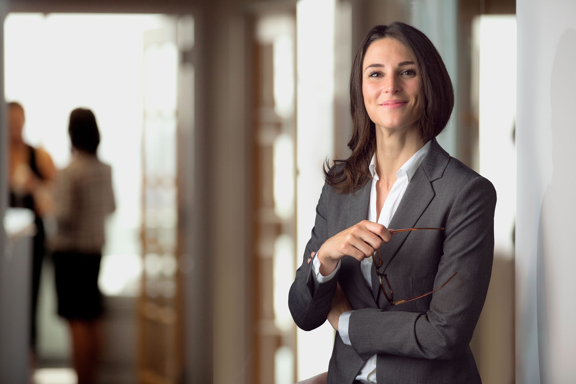 Woman in a business suit smiles, holding glasses, standing in a hallway; two figures are blurred in the background.