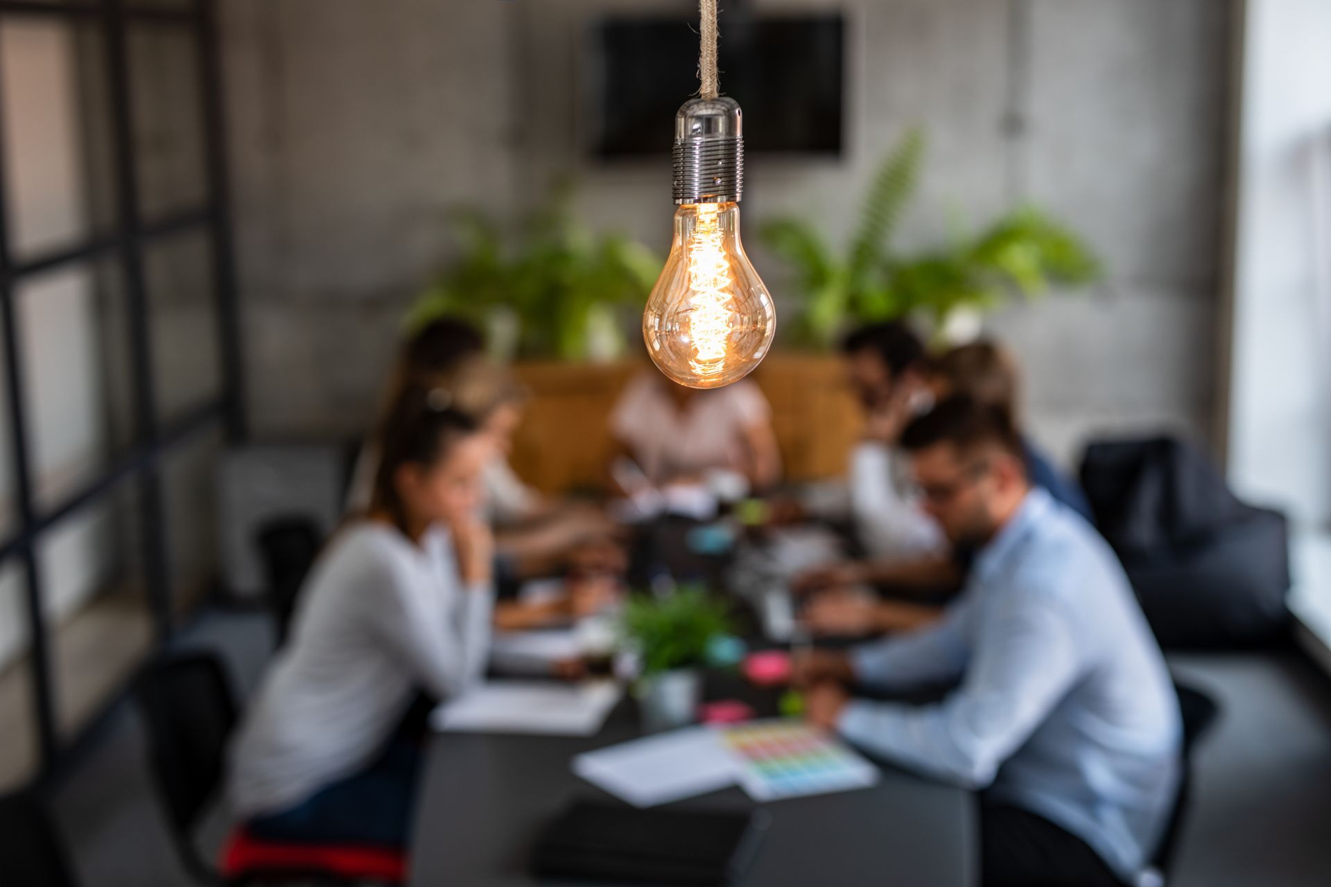 Lit bulb hangs above blurred office meeting.