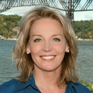 Woman with blonde hair smiles, standing in front of a bridge and water, wearing a blue shirt.