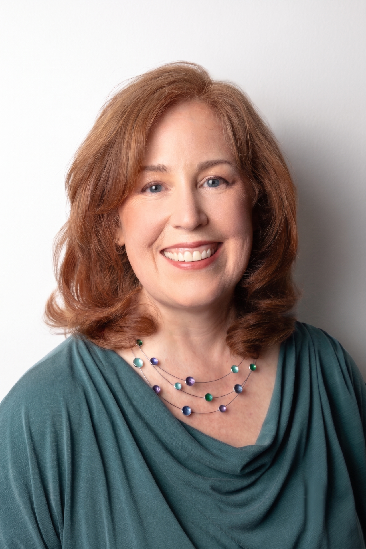 Woman with red hair, smiling in a blue top, arms crossed, leaning against a red brick wall.