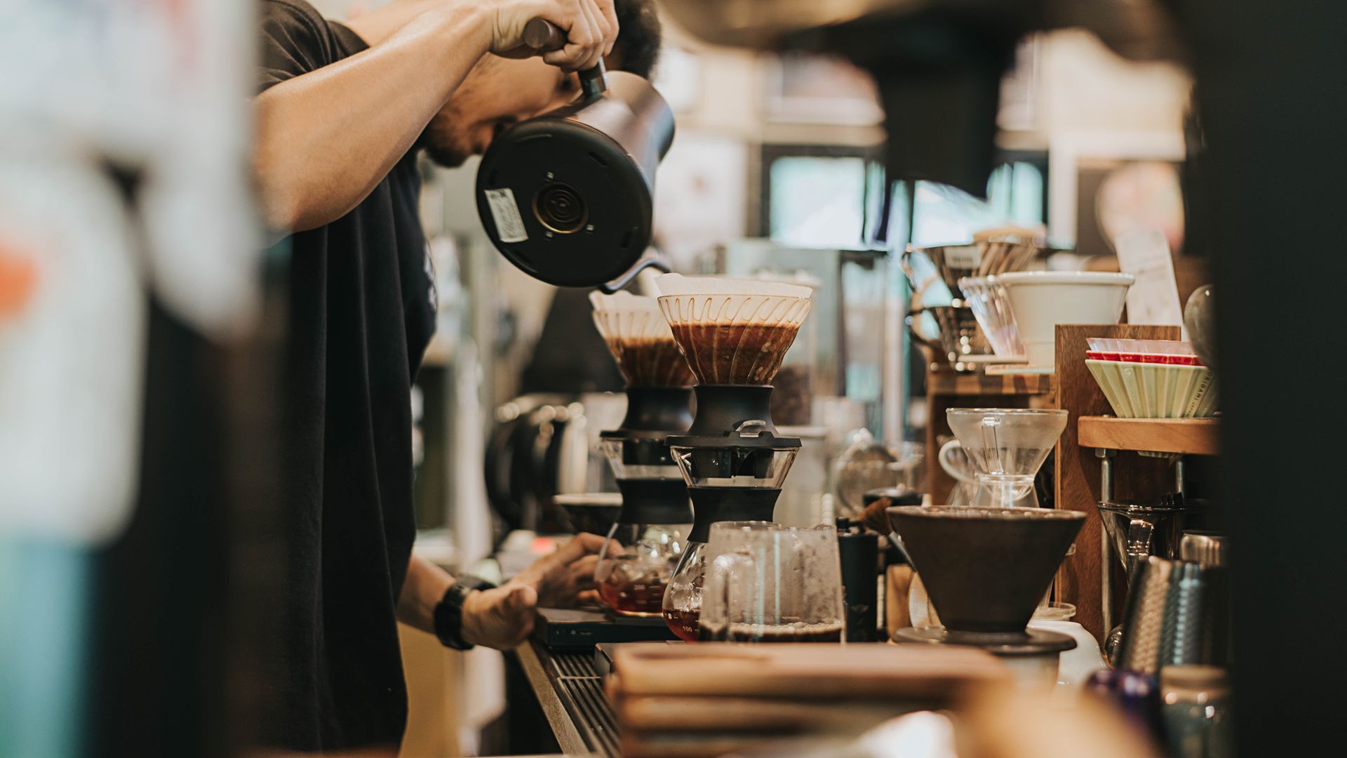 Barista pouring hot water into a coffee dripper at a café counter, with brewing equipment in view