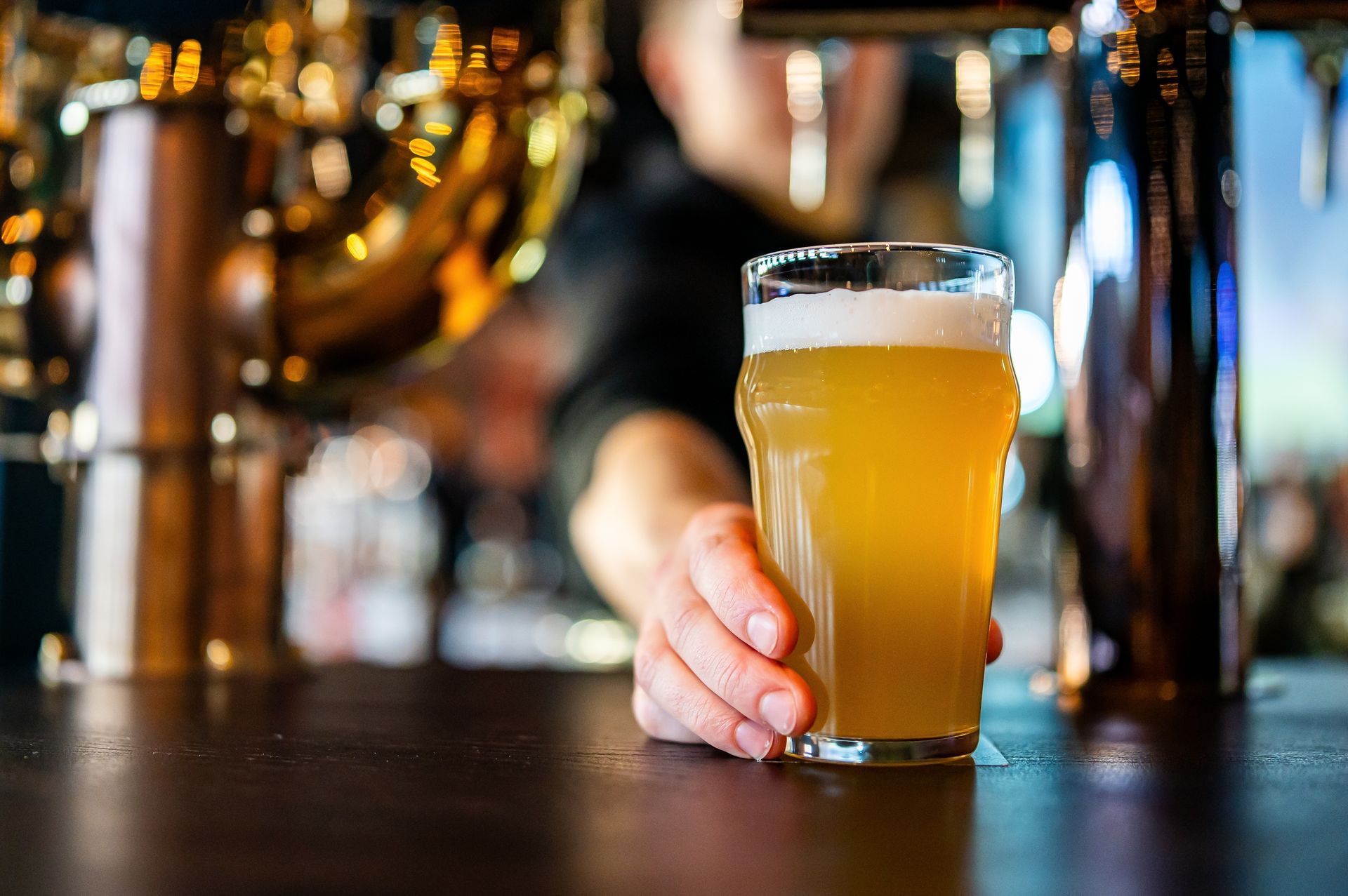 Hand holding a foamy pint of beer on a bar counter with blurred glasses in the background