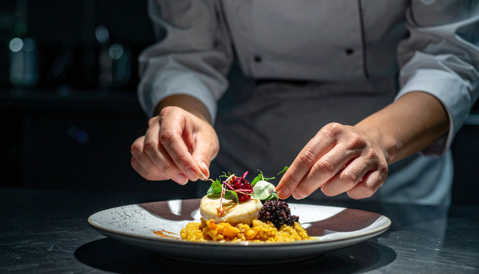 Chef plating a gourmet dish with garnishes on a white plate in a dim kitchen