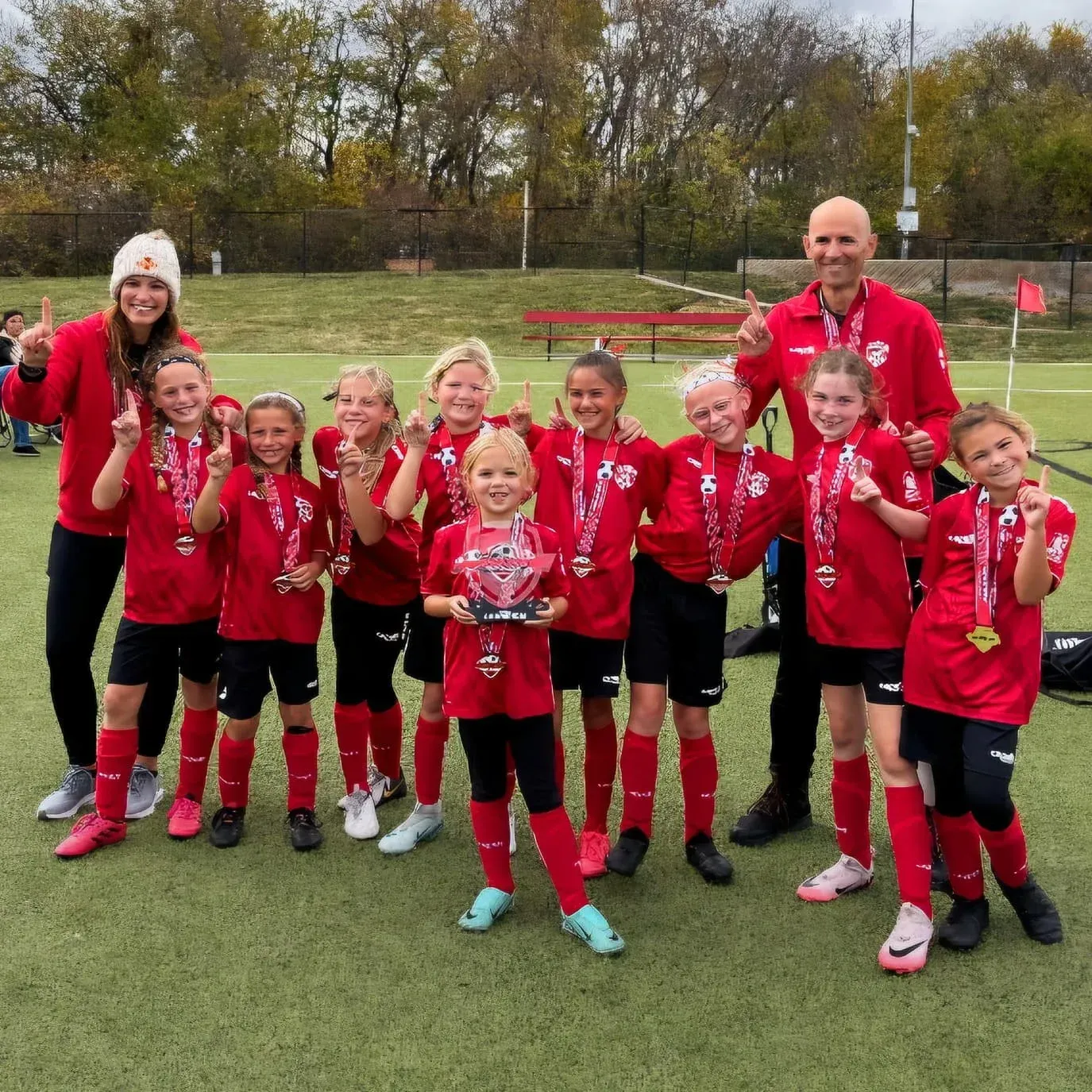 Youth soccer team and coaches posing with medals on a field, wearing red jerseys.
