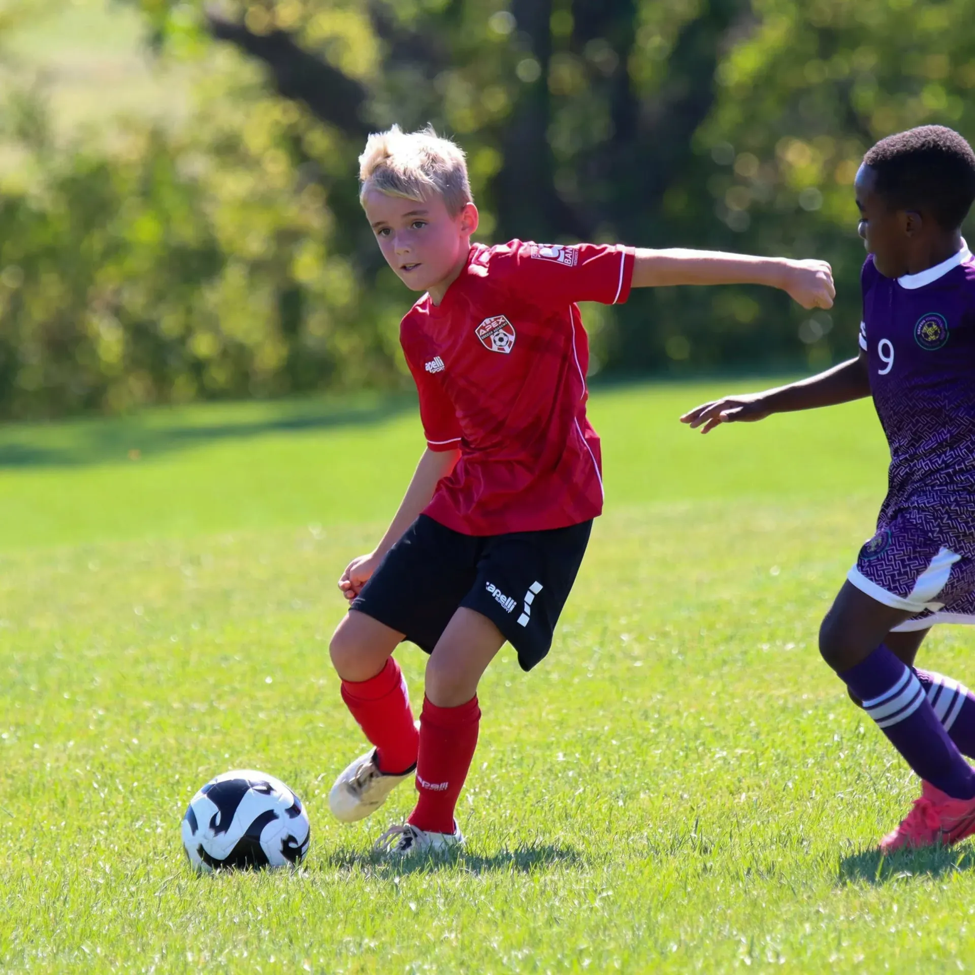 Two children playing soccer on a sunny grassy field, one in a red jersey dribbling the ball