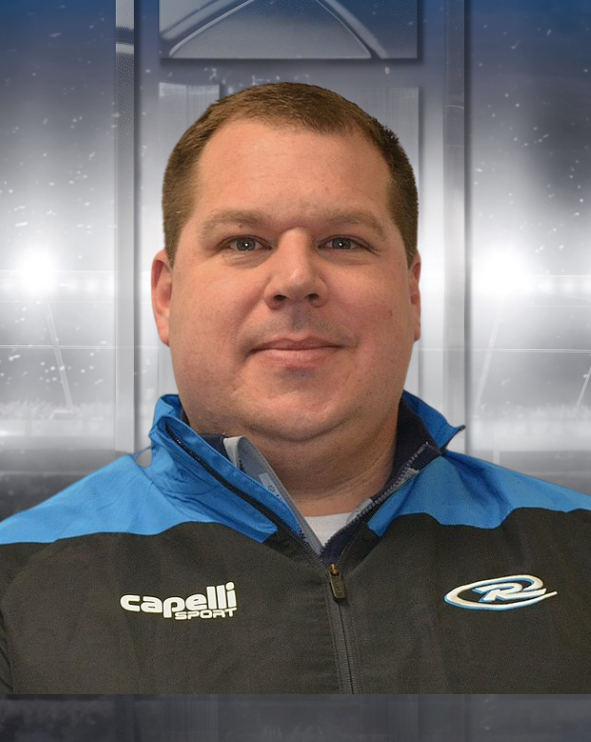 Man wearing a black-and-blue jacket, standing before a softly lit indoor sports backdrop.