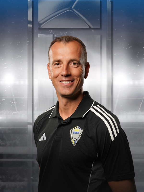 Man in black Adidas polo with team crest, smiling against a stadium-like gray backdrop
