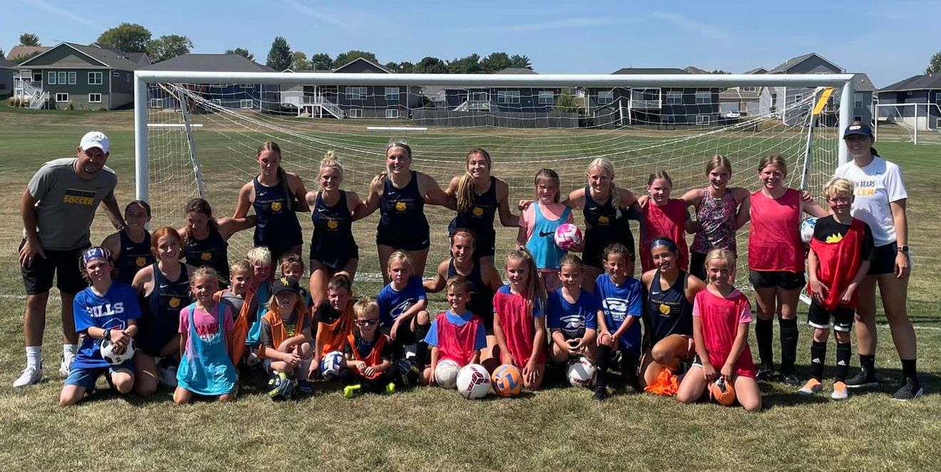 Youth soccer team posing in front of a goal on a grassy field, wearing blue and red uniforms.