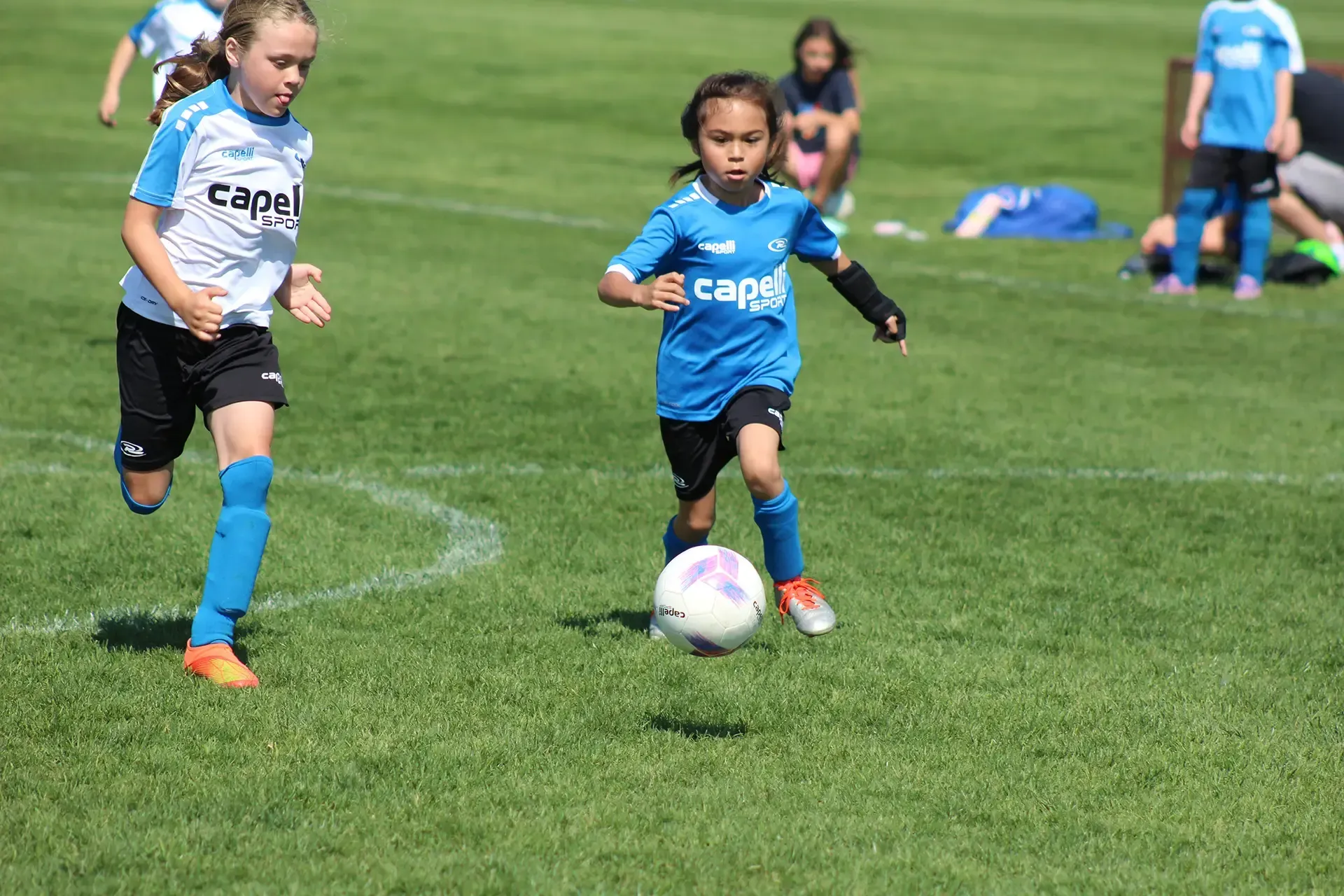 Two children playing soccer on a grassy field, with the ball at the center and teammates in the background