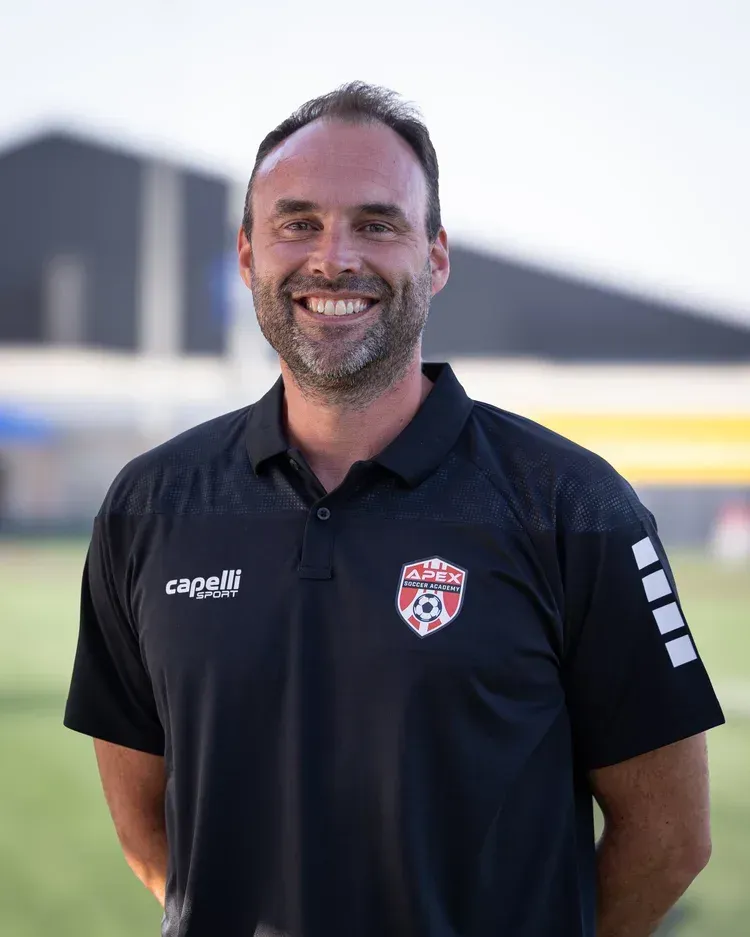 Smiling man in a black polo shirt standing outdoors with a blurred building in the background
