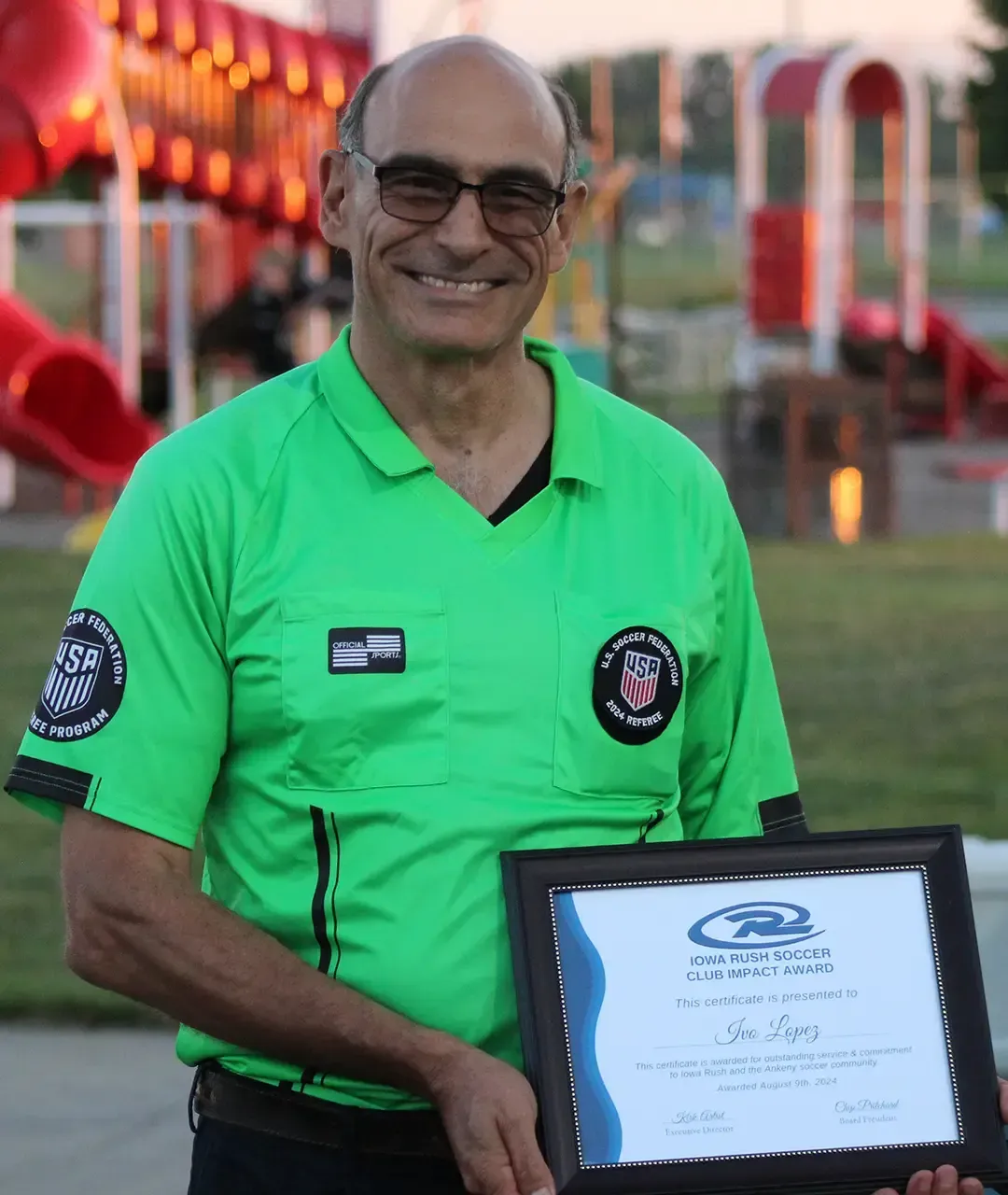 Smiling man in a bright green shirt holding a framed certificate outdoors at an event