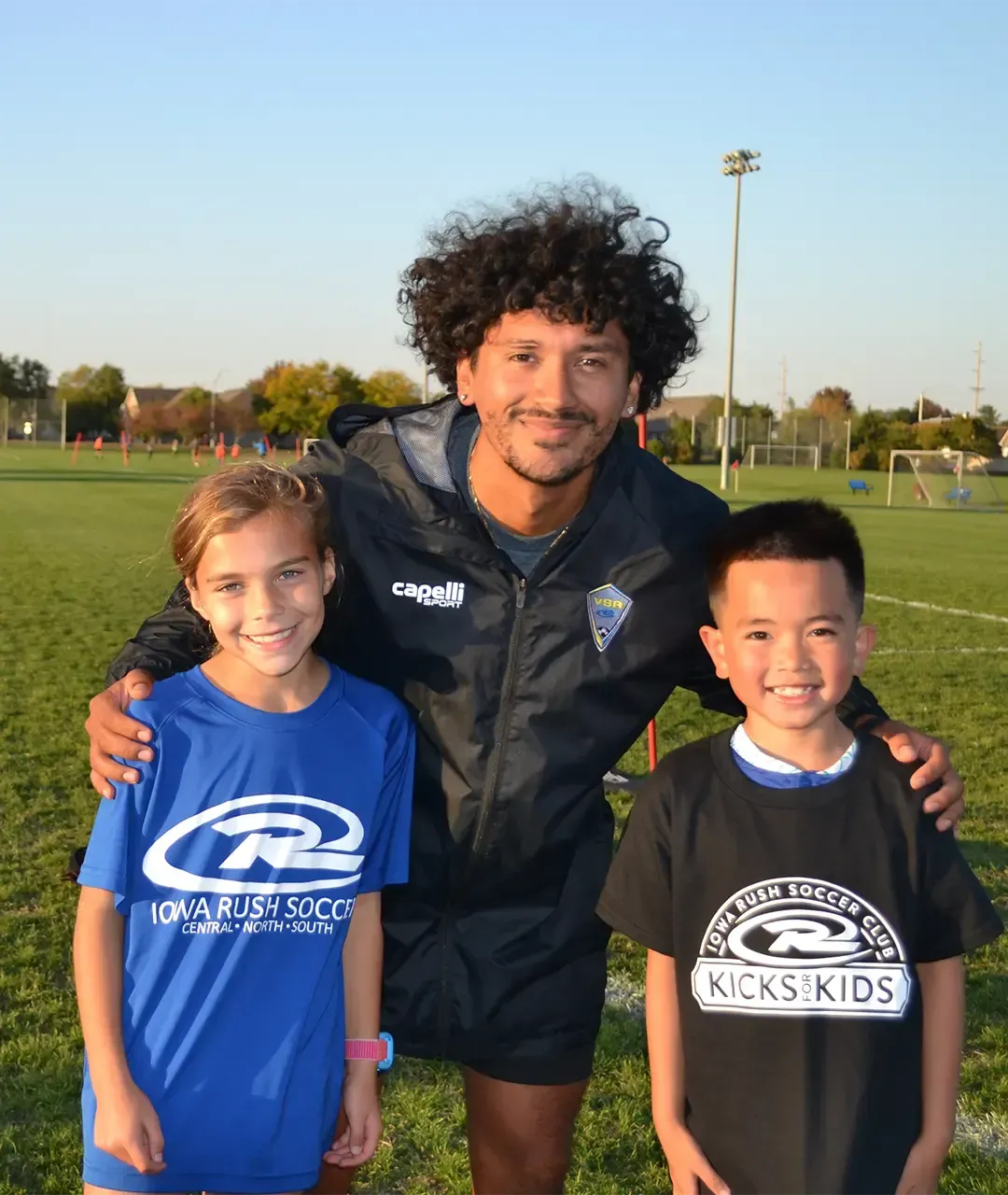 Three people smiling on a grassy sports field, with a man standing between two children