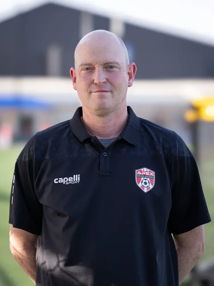 Man in black polo shirt with a soccer crest, standing outdoors against a blurred building background