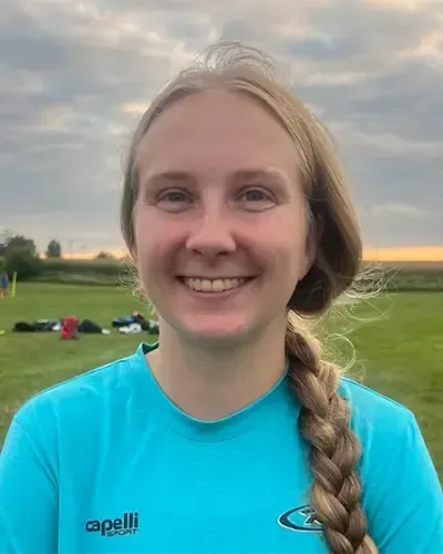 Smiling athlete in a turquoise shirt with a braid, standing on a grassy field at sunset.