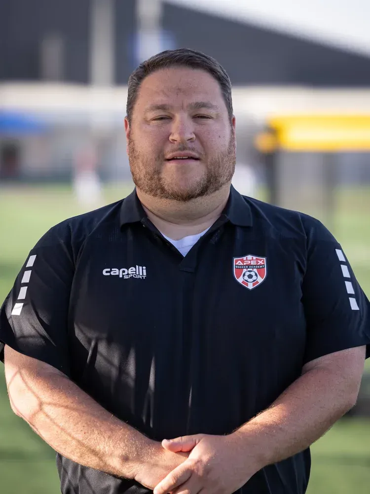 Man in black sports shirt standing outdoors, wearing a team crest, hands clasped, with a blurred background.