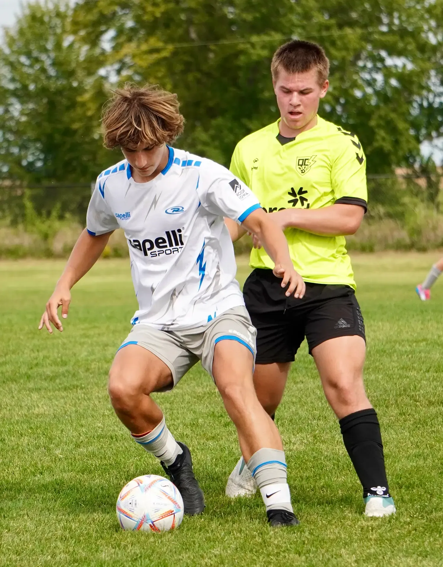 Two soccer players contesting the ball on a grassy field, one in white and blue, one in neon yellow.