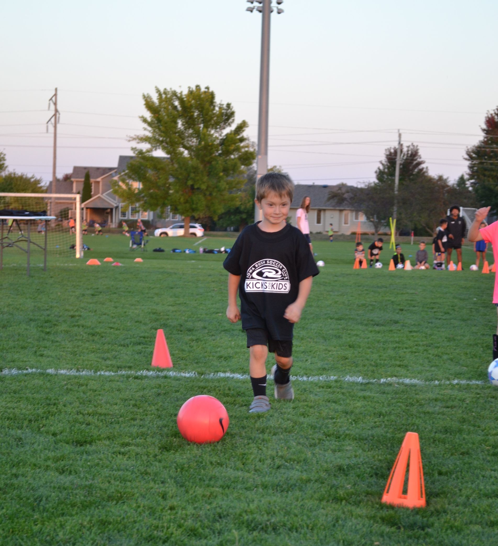 Child on a soccer field dribbling a red ball between orange cones at dusk.