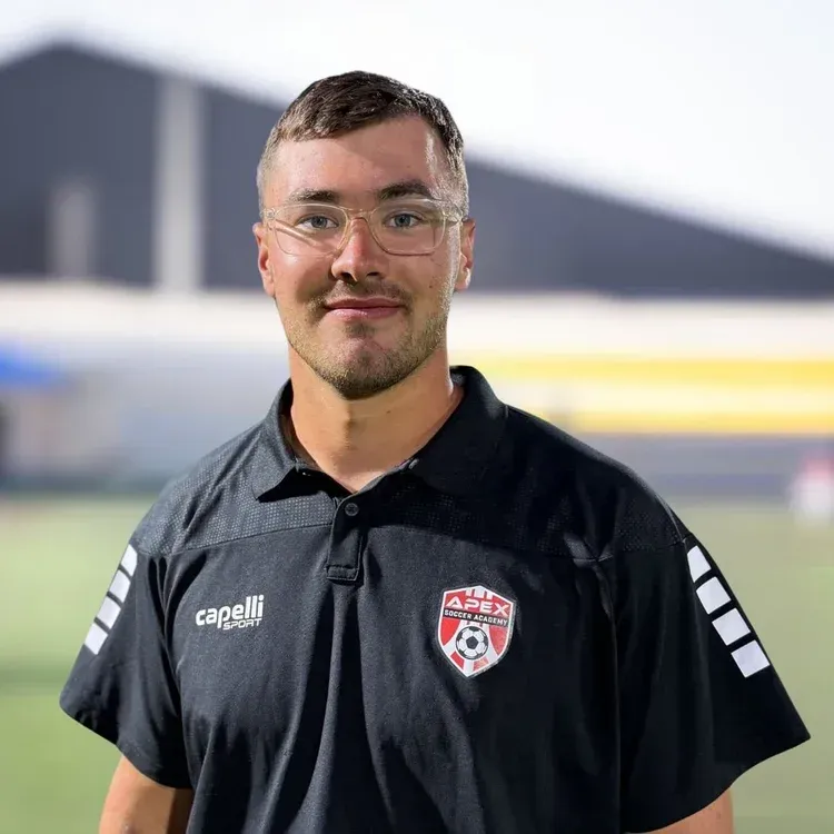 Portrait of a man in a black polo shirt with a club crest, standing on a blurred sports field background