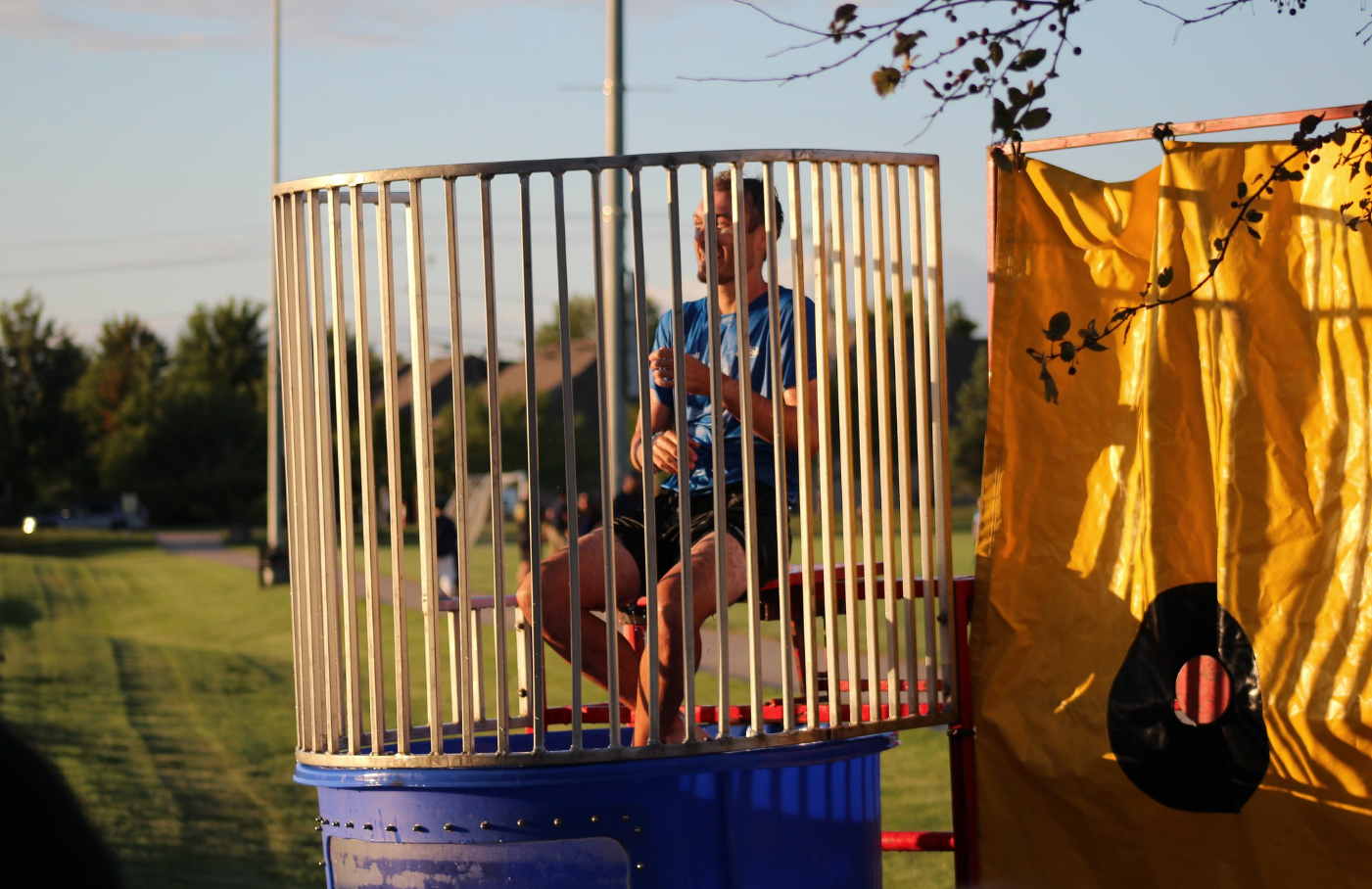 Person seated in a dunk tank at an outdoor event, with blue barrel and yellow target panel.