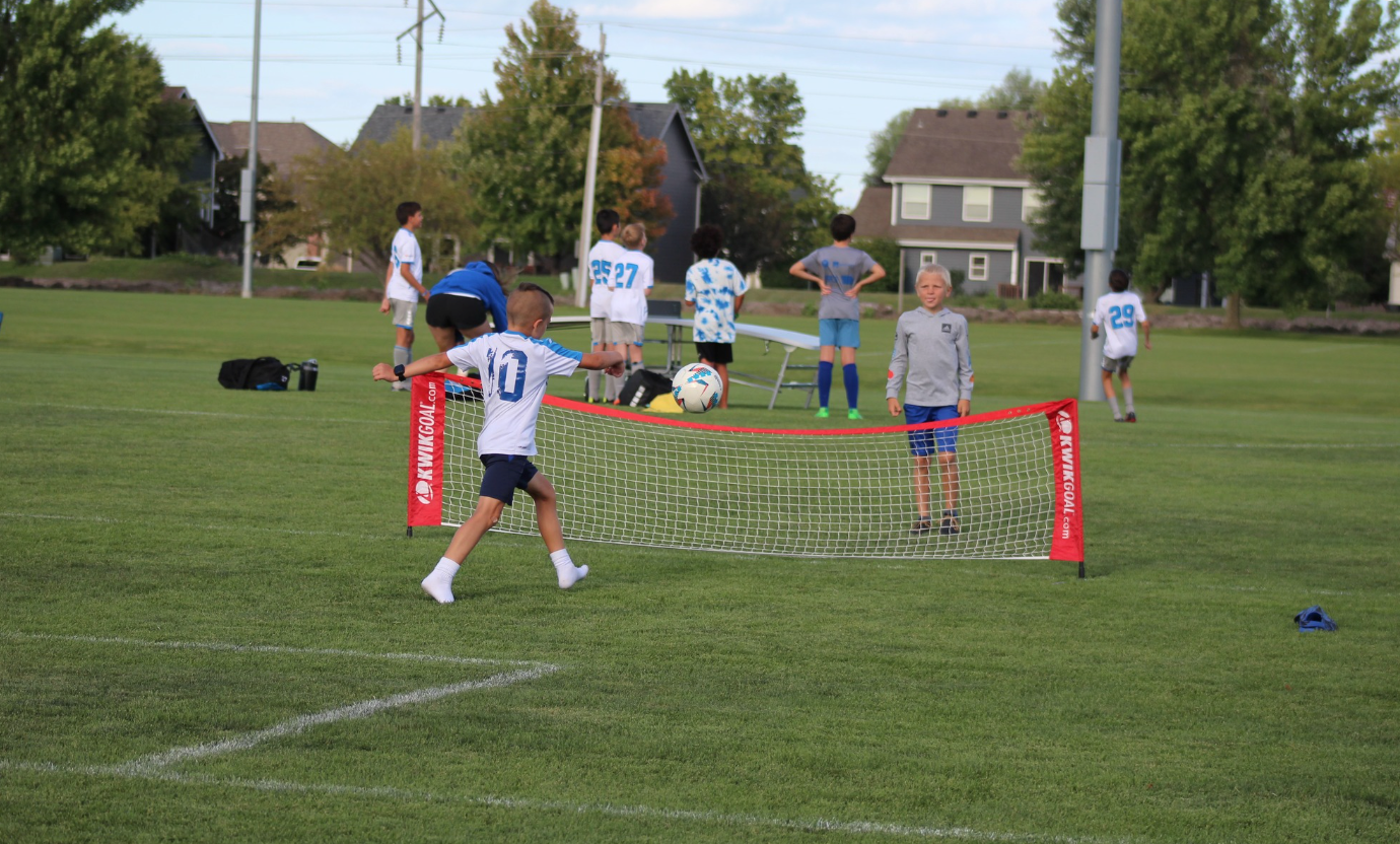 Children playing soccer on a grassy field with a small red goal and a net barrier in front.