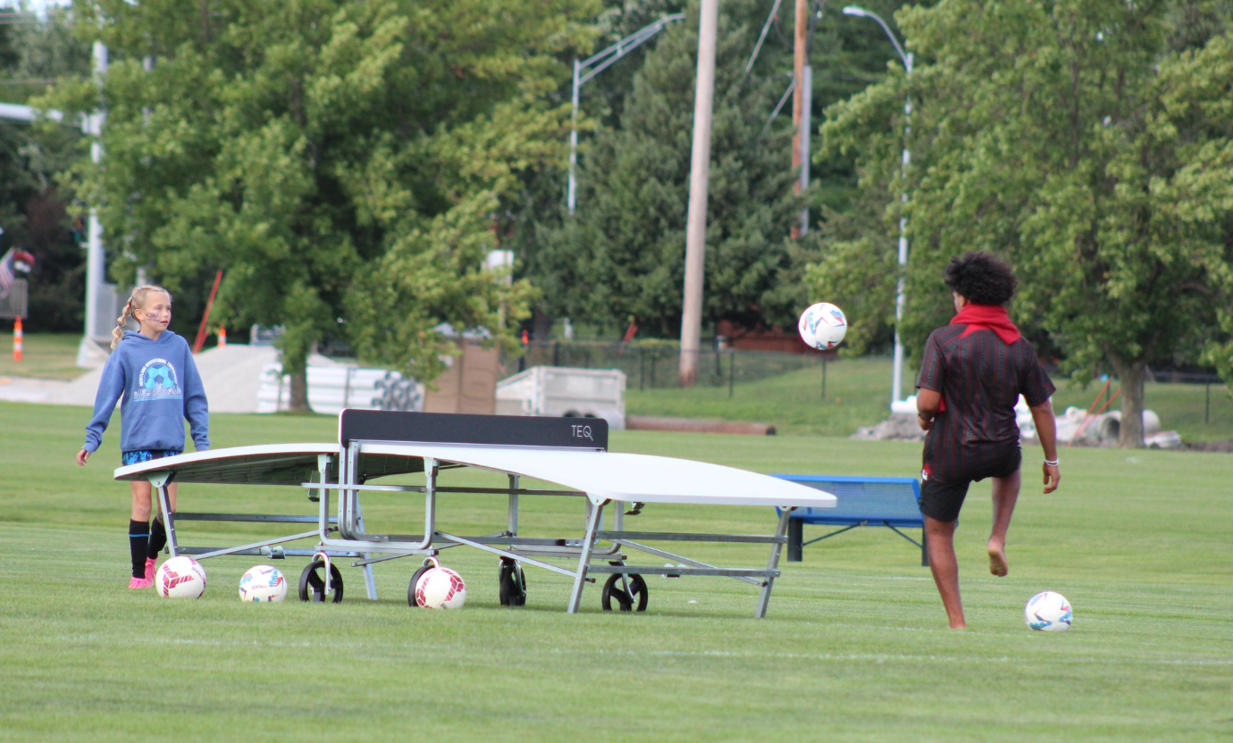 Two people on a grassy field near a ping-pong table, with soccer balls scattered around.