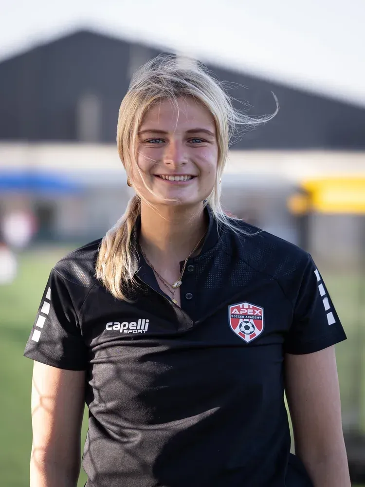 Smiling athlete in a black polo shirt standing outdoors in front of a blurred field and buildings