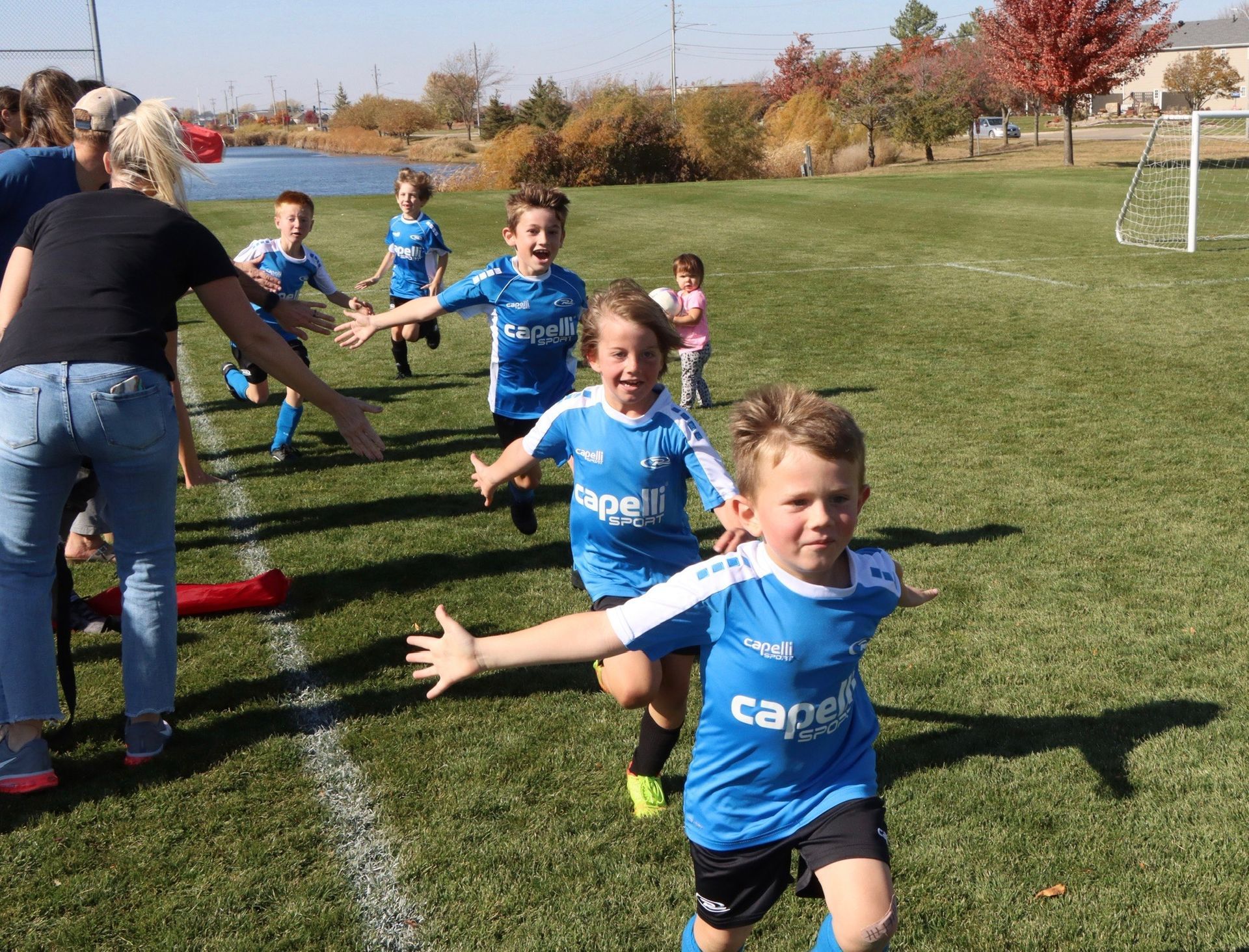 Children in blue soccer uniforms running on a grassy field toward the camera and a coach at left