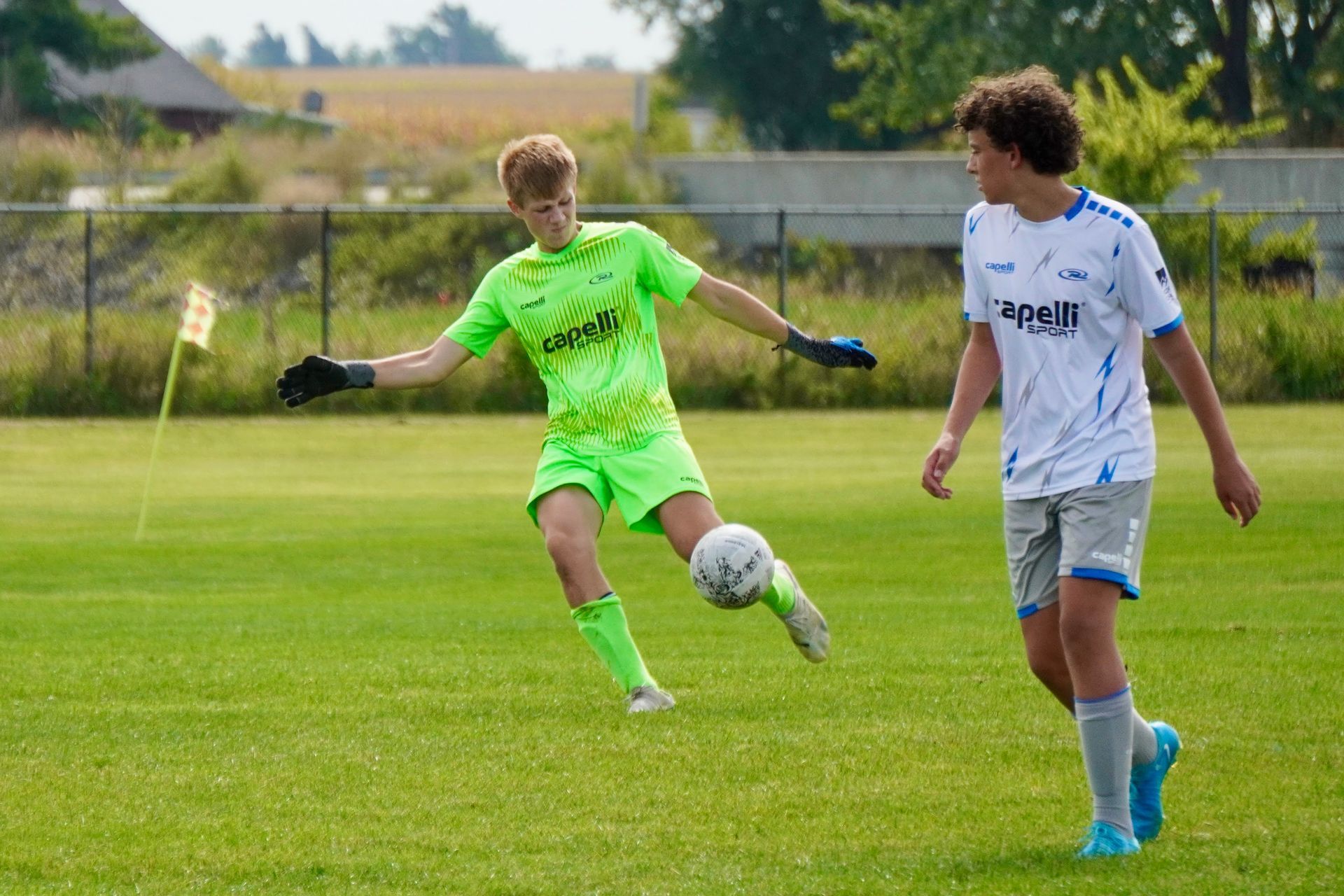 Two soccer players contest a ball on a grassy field, one in neon green kicking and one in white watching.