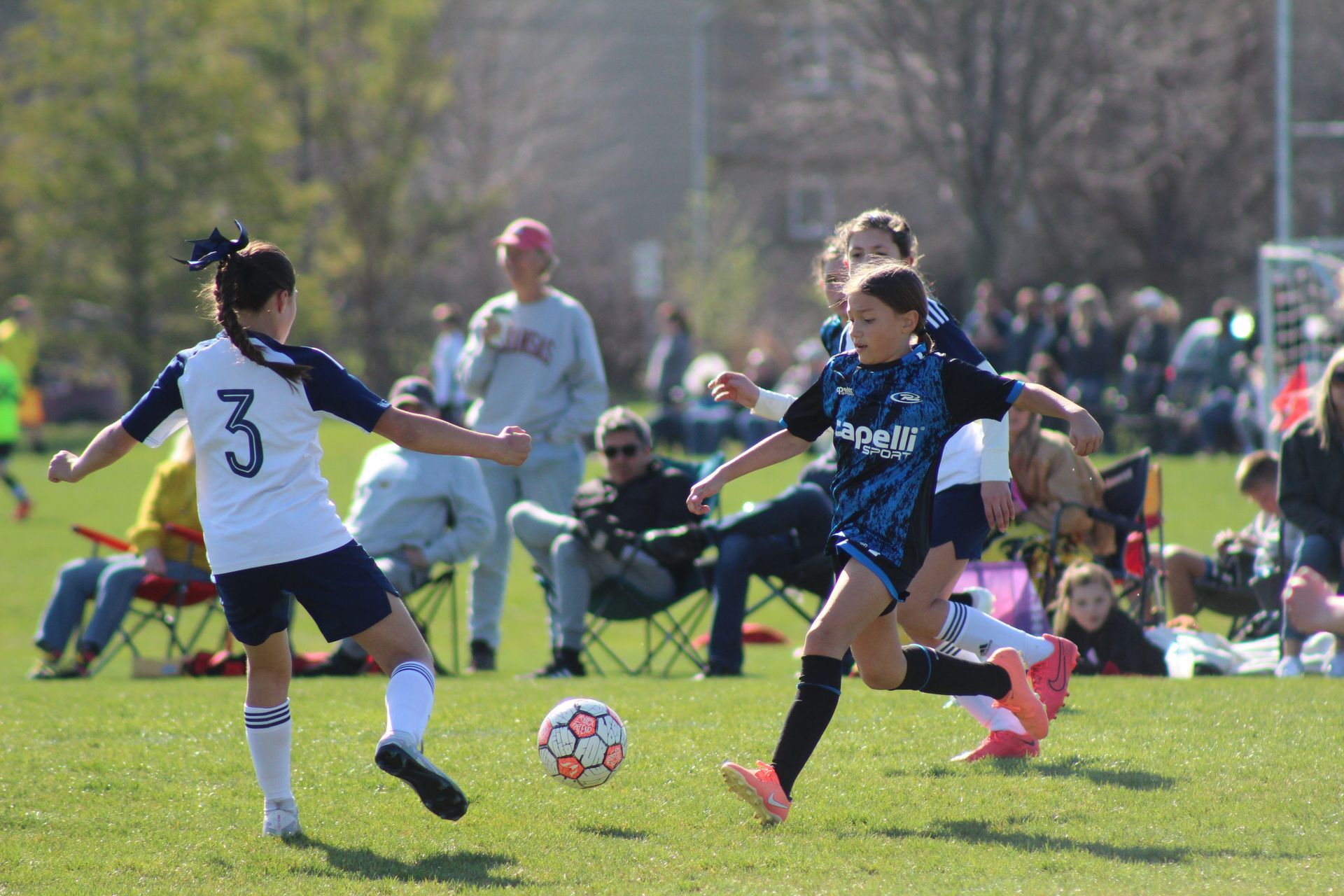 Two soccer players in white and blue jerseys chase the ball on a grassy field during a match.