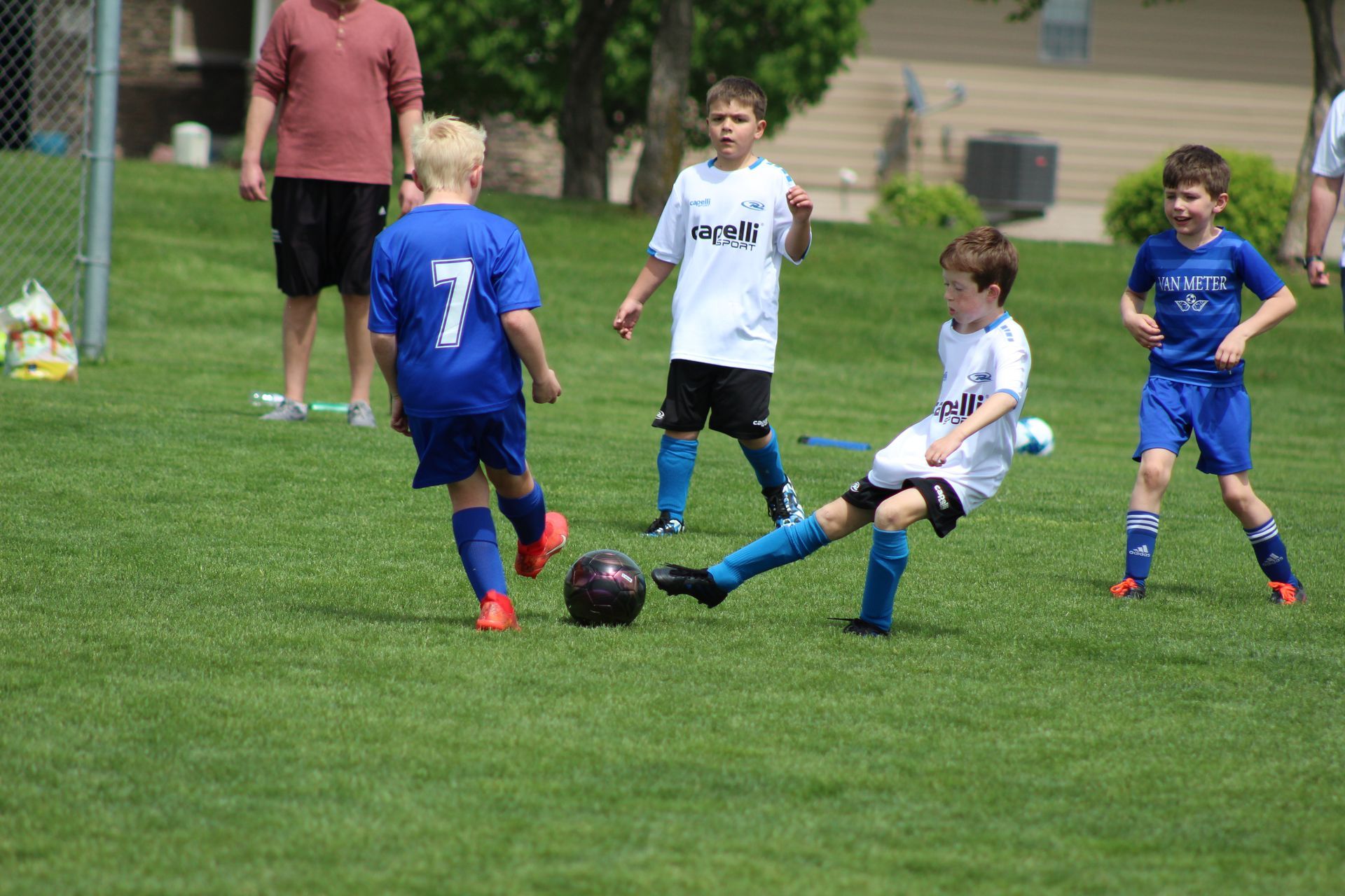 Children playing soccer on a grassy field, one in white kicking the ball as others in blue watch.