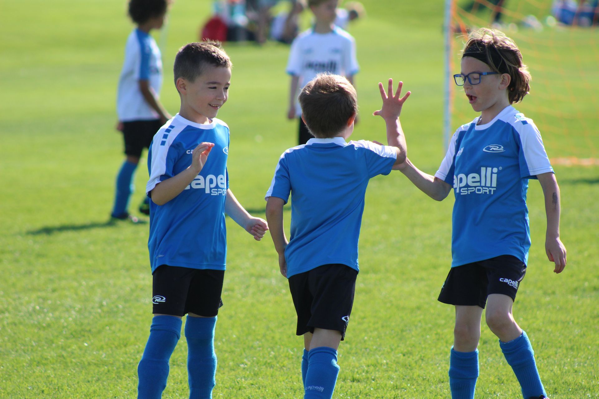Three children in blue soccer uniforms stand on a grassy field, one raising a hand for a high-five.
