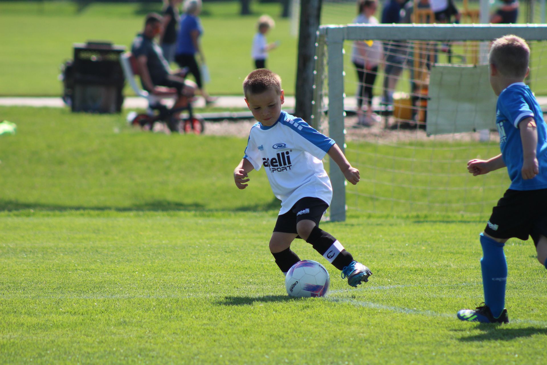 Two children playing soccer on a sunny grassy field, one dribbling the ball near the goal.