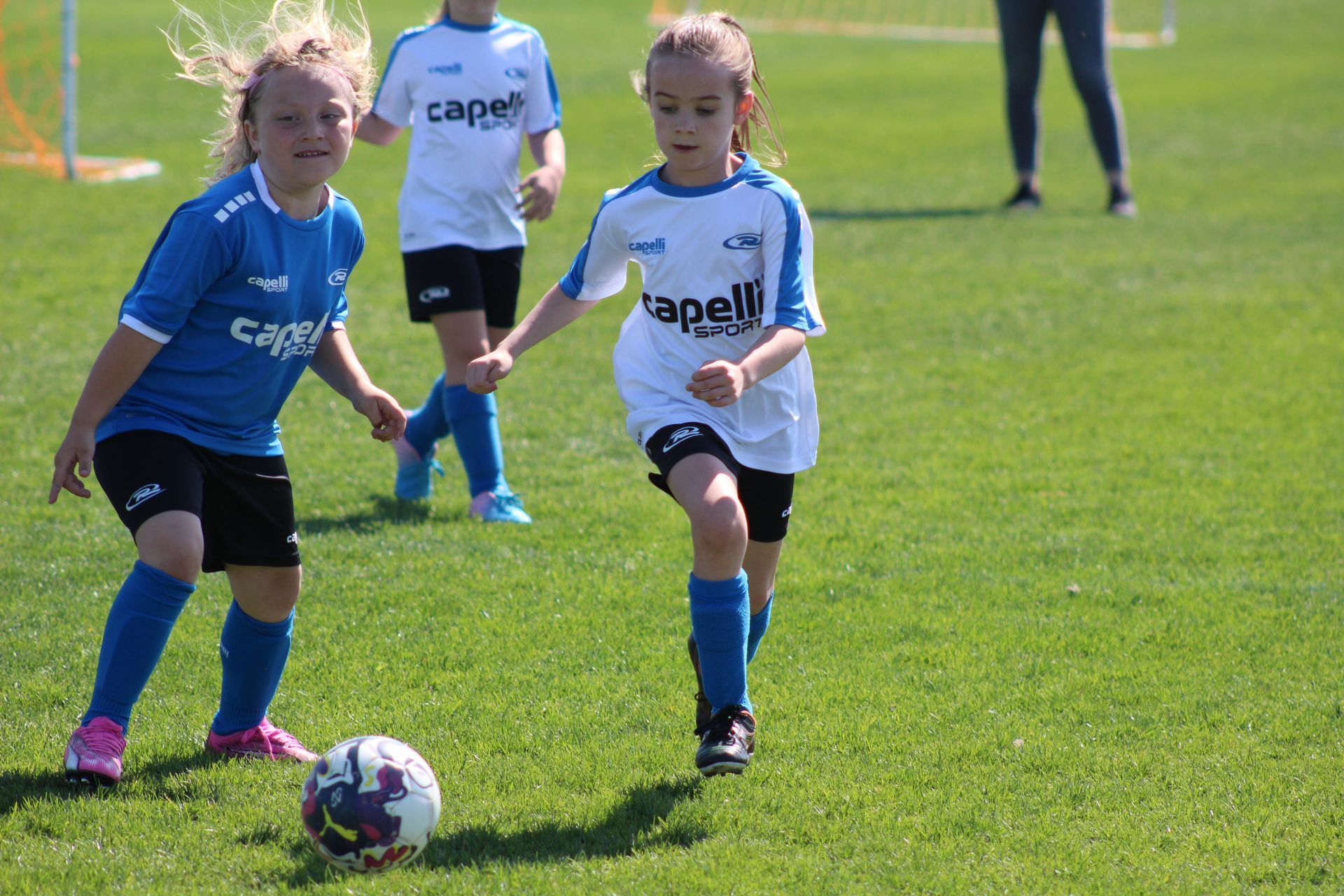 Two children in blue soccer uniforms chasing a ball on a grassy field