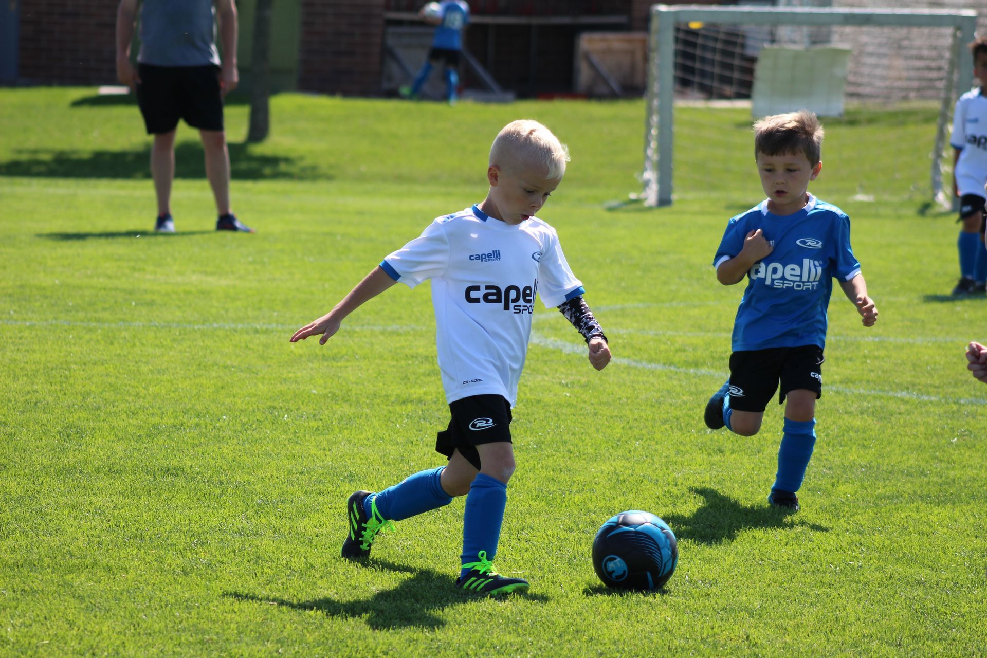 Two children in soccer uniforms chasing a ball on a grassy field near a goal