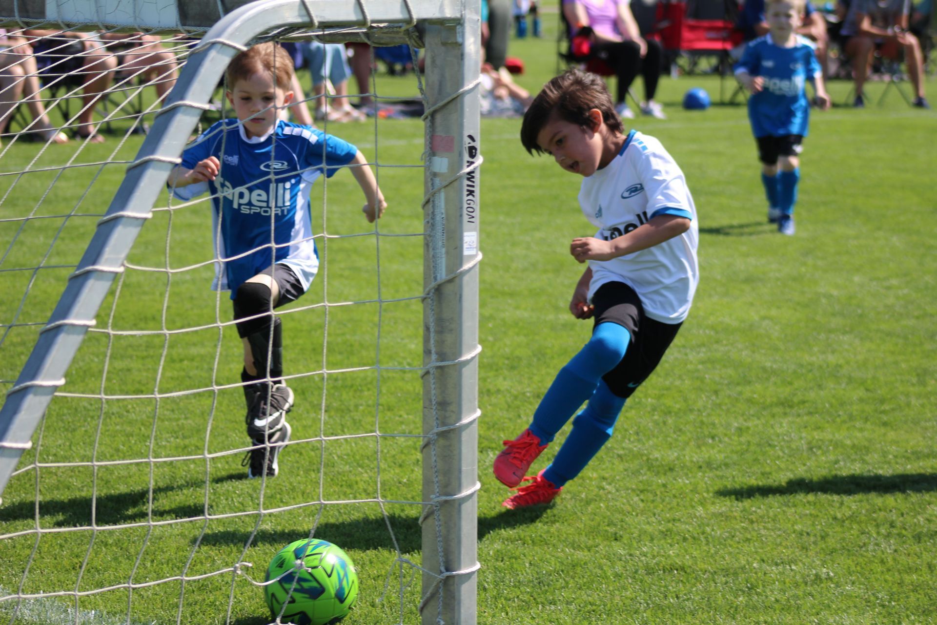 Two children playing soccer, one in blue by the goal and one in white kicking a green ball on the field