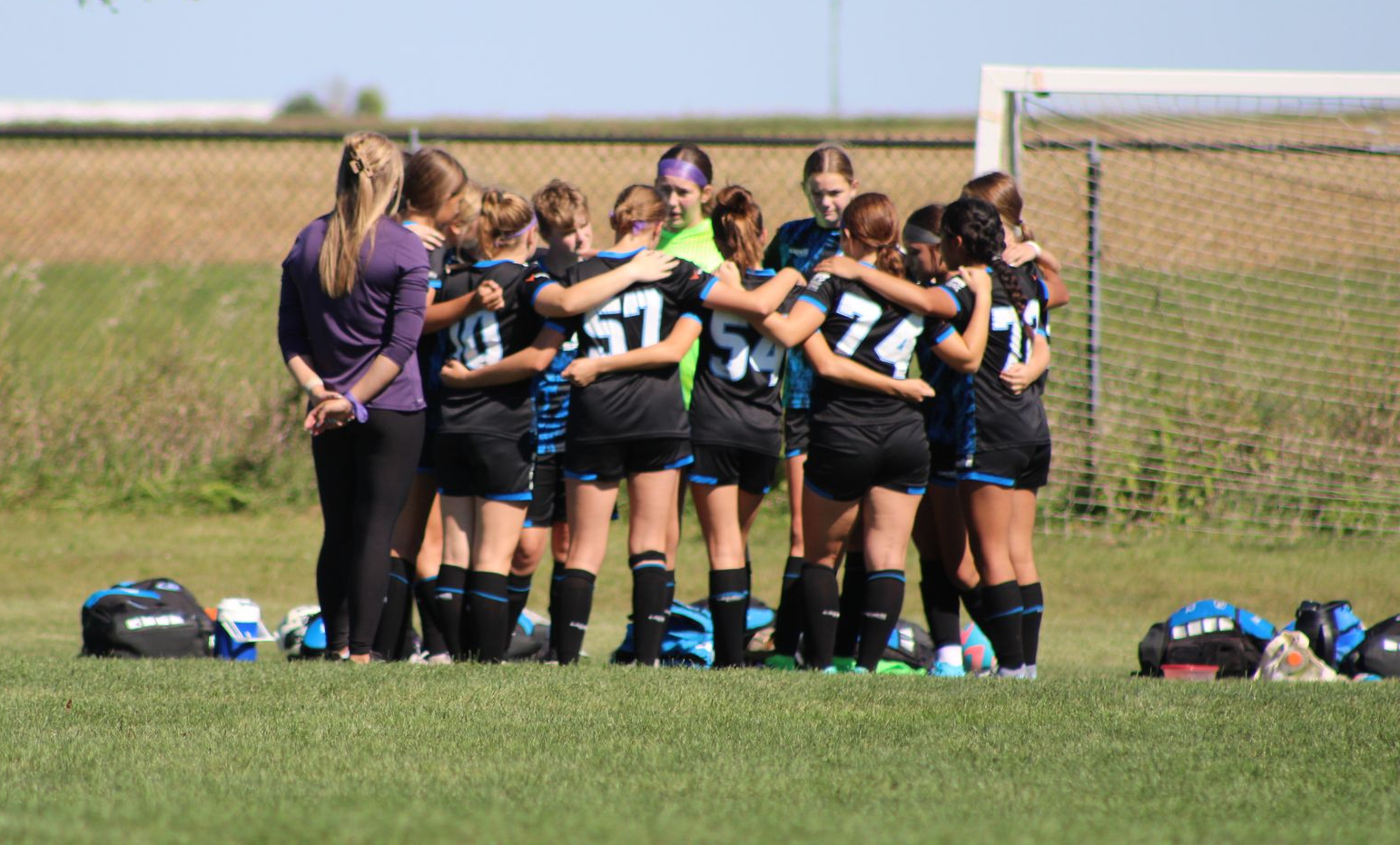 Women’s rugby team huddling on a grassy field, coaches nearby, before a match