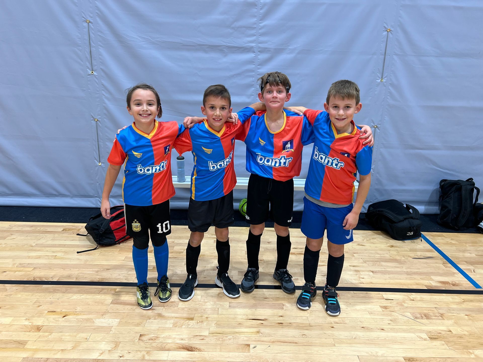 Four young soccer players in red and blue uniforms posing on an indoor court