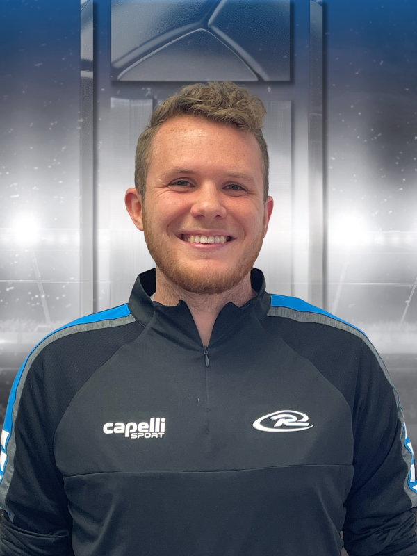 Smiling person in a black sports jacket with blue stripes, standing in front of a gray industrial background