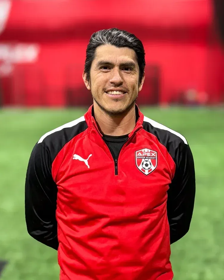 Soccer player in red and black training jacket standing on a field, smiling at the camera
