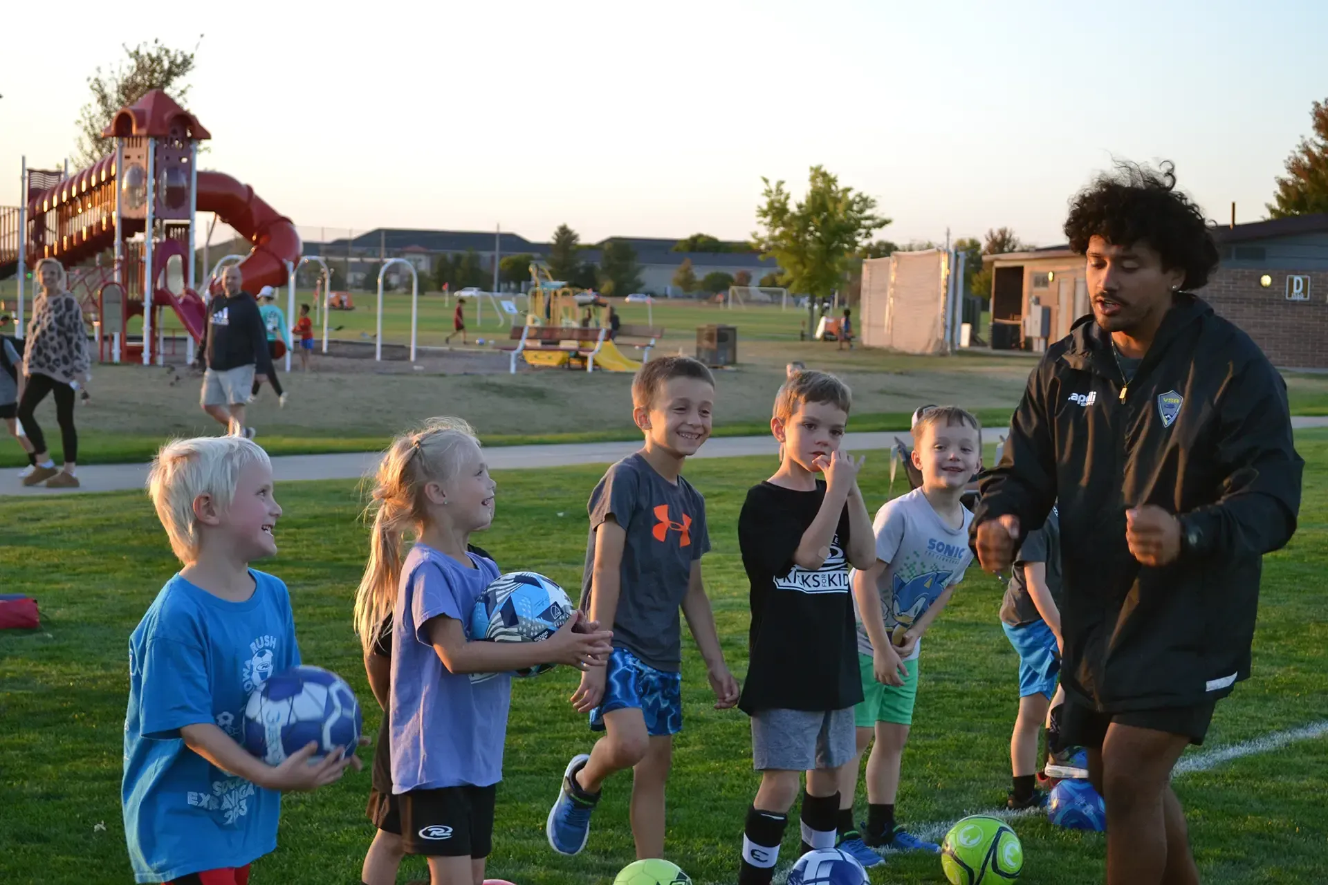 Children gathered with a coach on a soccer field at sunset, holding balls and wearing cleats.